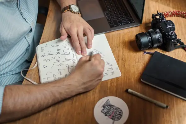 A man drawing logo designs in a sketchbook on a desk with a laptop, camera and notebook nearby